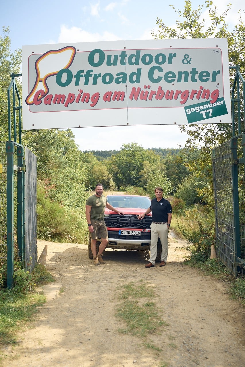Two men pose with a truck beneath a sign that reads "Outdoor & Offroad Center Camping am Nurburgring gegenuber T7"